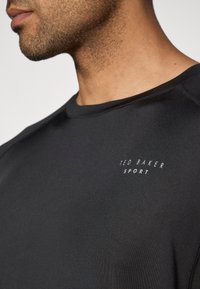 Juoda sportinė marškinė su apvaliu iškirpimu. Viršutiniame kairiajame kampe yra tekstas "TED BAKER SPORT" balta spalva. Lygus audinio paviršius.