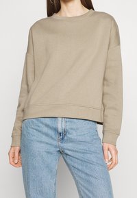 Sweat-shirt marron clair avec un col rond et des épaules tombantes. Comprend des poignets et un ourlet côtelés, fabriqué à partir d'un tissu doux et texturé.