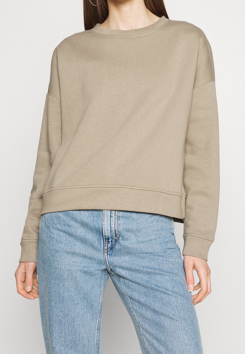 Sweat-shirt marron clair avec un col rond et des épaules tombantes. Comprend des poignets et un ourlet côtelés, fabriqué à partir d'un tissu doux et texturé.