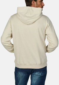 Beige hoodie with a drawstring hood, soft texture, ribbed cuffs, and hem. Worn over dark blue jeans, showing a relaxed fit.