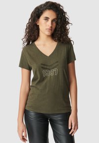 Camiseta verde oliva de cuello en V con un estampado texturizado que dice "1987" en blanco. Mangas cortas, tejido suave y corte casual. Pantalones de cuero negros.