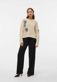 Pull en tricot beige avec broderie florale noire, associé à un pantalon large noir. Ce look présente une coupe décontractée et des poignets côtelés.