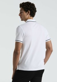 Weißes Polohemd mit schwarzen Akzenten am Kragen und an den Ärmeln, klassischer Schnitt und glatte Textur, getragen mit schwarzen Hosen.