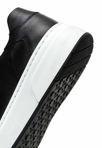 Zapatilla negra de cuero con suela de goma blanca. Presenta una suela texturizada negra con un patrón geométrico. Parte superior suave con costuras mínimas.