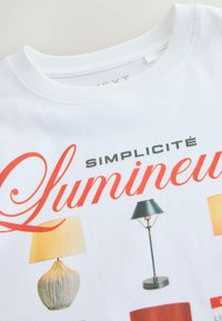 Tricou alb din bumbac cu un design imprimat ce prezintă lămpi în diverse stiluri și culori, însoțit de textul „SIMPLICITÉ LUMINEUSE” în roșu.