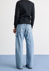Light blue denim wide-leg jeans with a relaxed fit, featuring rear pockets and a loop accent. Paired with a black sweatshirt.