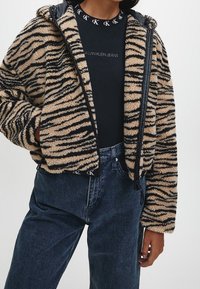 Kurtka z futra sztucznego w paski w kolorze tan i czarnym, zapinana na zamek z kapturem, noszona na czarnej koszulce Calvin Klein i zestawiona z ciemnymi dżinsami.