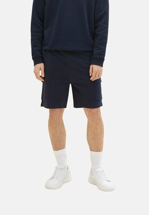 Uomo che indossa pantaloni cargo blu navy, una maglietta a maniche lunghe blu navy, calze bianche a girocollo e sneakers bianche in piedi contro uno sfondo neutro.