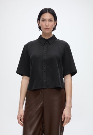 Žena s krátkými černými vlasy, oblečená v volné černé košili na knoflíky a hnědých kožených kalhotách, stojící před jednobarevným bílým pozadím.