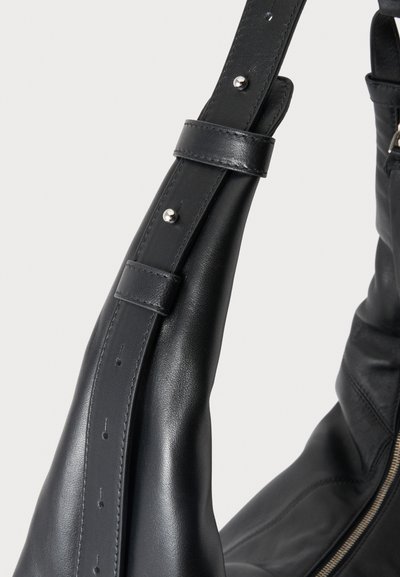 Gros plan sur une bandoulière de sac en cuir noir dotée de clous métalliques réglables et de boucles, à côté de la fermeture éclair dorée du sac.