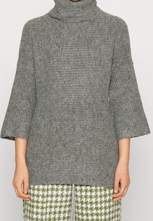 Pull gris en maille à col roulé avec manches larges trois-quarts, porté avec un pantalon pied-de-poule vert et blanc, mains détendues le long du corps.