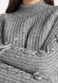 Grauer gestrickter Pullover mit hohem Kragen, ausgestattet mit geripptem Textil und dekorativen geflochtenen Details entlang der Ärmel.