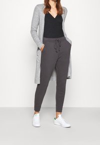 Harmaa neuletakki, musta V-kauluksinen paita, tummanharmaat nyörillä varustetut collegehousut ja valkoiset lenkkarit, joissa vihreät yksityiskohdat. Rentoon ja mukavaan tyyliin sopiva asu.