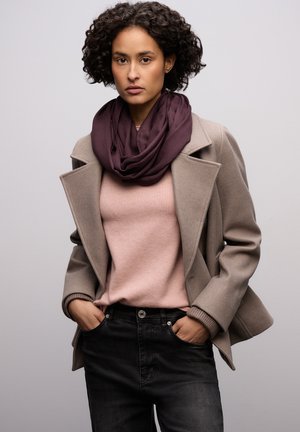 Beige jas over een roze trui, zwarte spijkerbroek. Een donkerpaarse sjaal hangt om de nek. Het model heeft krullend haar en de handen in de zakken.