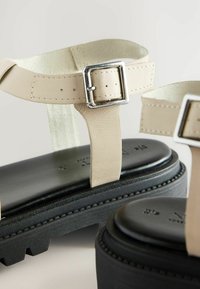 Beige bőr szandálok állítható ezüst csattal, vastag fekete platform talppal és texturált pántokkal. Simább felület és varrott részletek.