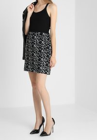 Woman wearing black tank top, leopard print mini skirt with zipper, holding black jacket, and black high-heeled shoes against white background.