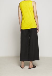 Keltainen ilman hiuksia oleva paita, jossa on tekstuuri, yhdistettynä leveälahkeisiin mustiin housuihin ja mustiin sandaaleihin, nähtynä takaa.