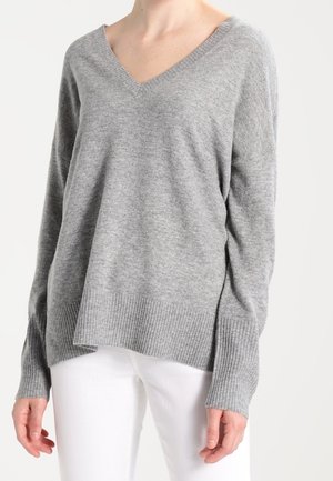 Femme portant un pull ample gris clair à col en V avec des poignets et un ourlet côtelés, associé à un pantalon blanc.