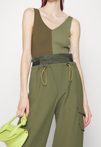 Top a canotta a coste verde oliva con scollatura a V, abbinato a pantaloni larghi coordinati. L'outfit presenta una cintura a contrasto e una piccola borsa verde.