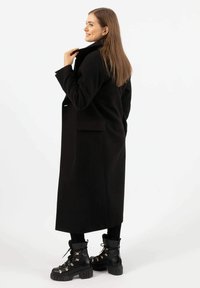 Zwarte wollen jas met een klassiek snit, zijzakken en een kraag, gecombineerd met zwarte veterschoenen met gouden accenten en gestructureerde zolen.