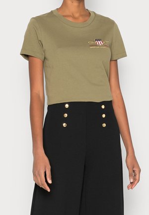 Persoon die een olijfgroen T-shirt met korte mouwen draagt met een klein logo en een zwarte broek met hoge taille met twee rijen gouden knopen.