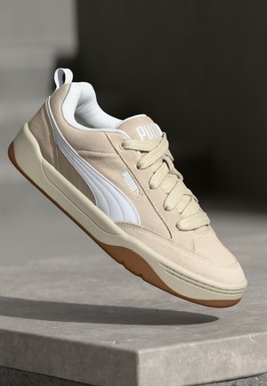 Zapatilla beige con una franja lateral blanca, parte superior texturizada, cordones tonales y suela de goma color caramelo, con una sutil marca en la lengüeta.