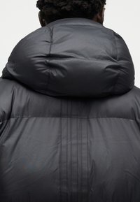 Άτομο με κοντά, σγουρά μαλλιά που φοράει μαύρο επενδυμένο μπουφάν με μεγάλη κουκούλα, φαίνεται από πίσω σε απλό φόντο.