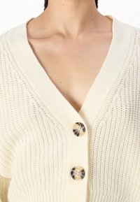 Krémový pletený cardigan s hlubokým V-výstřihem, třemi velkými texturovanými knoflíky a žebrovaným lemem. Měkký, texturovaný materiál.