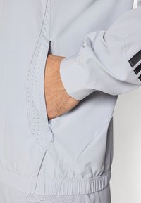 Ανοιχτό μπλε μπουφάν με φερμουάρ, που διαθέτει πλαϊνή τσέπη και διάτρητες λεπτομέρειες. Το ύφασμα φαίνεται λείο με ελαστικές μανσέτες και μέση.