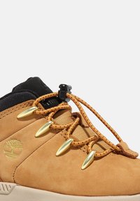 Chaussure en suede tan avec un col en tissu noir, dotée de quincaillerie dorée et de lacets élastiques. Logo d'arbre embossé sur le côté.