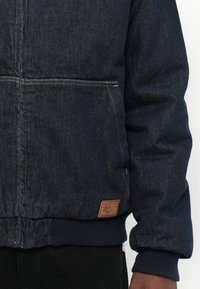 Temno modra denim jakna z rebrastim pasom in manšetami, s prsno žepom in majhnim rjavim usnjenim oznako na robu.