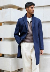 Abrigo de lana azul marino con cuello, superpuesto a un suéter marrón con cremallera y pantalones ligeros. El abrigo tiene un corte ajustado y bolsillos laterales.