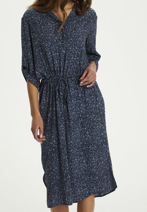 Frau trägt ein blaues Midikleid mit schwarzen abstrakten Mustern, dreiviertel Ärmeln und tailliert mit einem Kordelzug.