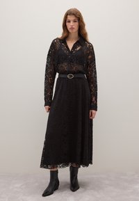 Vestido de encaje negro con mangas largas y cuello tipo camisa, combinado con un cinturón negro con una hebilla redonda. Usado con botas negras hasta el tobillo.