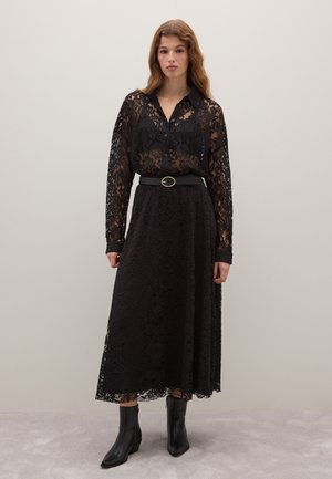 Vestido de encaje negro con mangas largas y cuello tipo camisa, combinado con un cinturón negro con una hebilla redonda. Usado con botas negras hasta el tobillo.