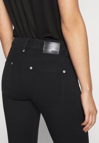 Vaqueros de mezclilla negra con una textura suave, que cuentan con una etiqueta de cuero con la marca en la parte trasera, dos bolsillos traseros y detalles metálicos en plata.