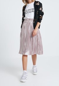  bomber jacket floreale nera sopra una t-shirt bianca con grafica, abbinata a una gonna plissettata metallica rosa e sneakers bianche.