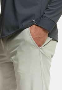 Graues Langarmoberteil mit glatter, matter Textur; hellfarbene Hose mit subtiler glänzender Oberfläche; Taschenbe detail am Hüftbereich sichtbar.