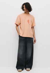 Tricou din bumbac de culoare piersică deschisă cu mâneci scurte, având un mic design de corali roșii pe buzunar. Asortat cu blugi lejeri, largi, de culoare gri închis.