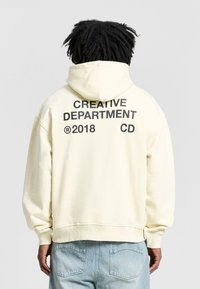 Kreemikas hoodie, millel on eesmine tasku ja seljal suured mustad kirjad: "KREATIIVNE DEPARTEME 2018 CD", pehme puuvillane materjal.