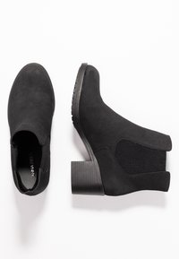 Un par de botas de tobillo de ante negro con paneles laterales elásticos y tacones cuadrados, mostradas desde vistas superior y lateral sobre un fondo blanco.