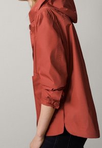 Person klädd i en rostorange huva med uppvikta ärmar, som står mot en enfaldig ljusgrå bakgrund.