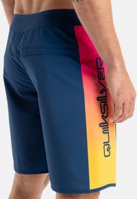 Granatowe szorty pływackie z gradientem różowym do żółtego na bocznym panelu, z tylną kieszenią i czarnym nadrukiem logo wzdłuż boku.