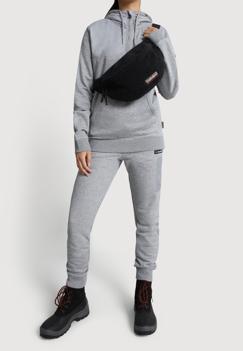 Szara bluza z kapturem, czarna torba, szare dresy i czarne buty. Torba ma miękką teksturę i widoczne logo marki.
