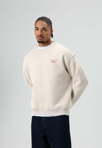 Beige sweatshirt met een ronde hals, geribbelde manchetten en zoom. Voorzien van een klein rood geborduurd logo op de borst. Gedragen over een wit shirt.