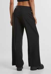 Pantaloni negri cu picioare largi, dintr-un material neted, cu talie înaltă și buzunare laterale discrete, asortați cu botine negre cu platformă groasă.