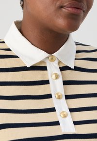 Top de punto a rayas beige y marina con cuello blanco y botones embossados en oro con forma de león a lo largo de la tapeta. Vista en primer plano del escote y los botones.