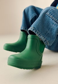 Stivali alla caviglia in gomma verde con suola testurizzata, design curvo e collo alto, indossati con pantaloni di denim blu. Superficie liscia senza motivi.