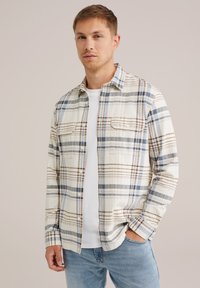 Plaid cotton shirt in cream with blue and brown stripes, buttoned front, two chest pockets, and a relaxed fit, layered over a white t-shirt.
