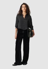 Camisa negra recortada de botones con mangas largas, combinada con pantalones anchos de terciopelo negros y un bolso de mano verde. Textura suave, diseño minimalista.